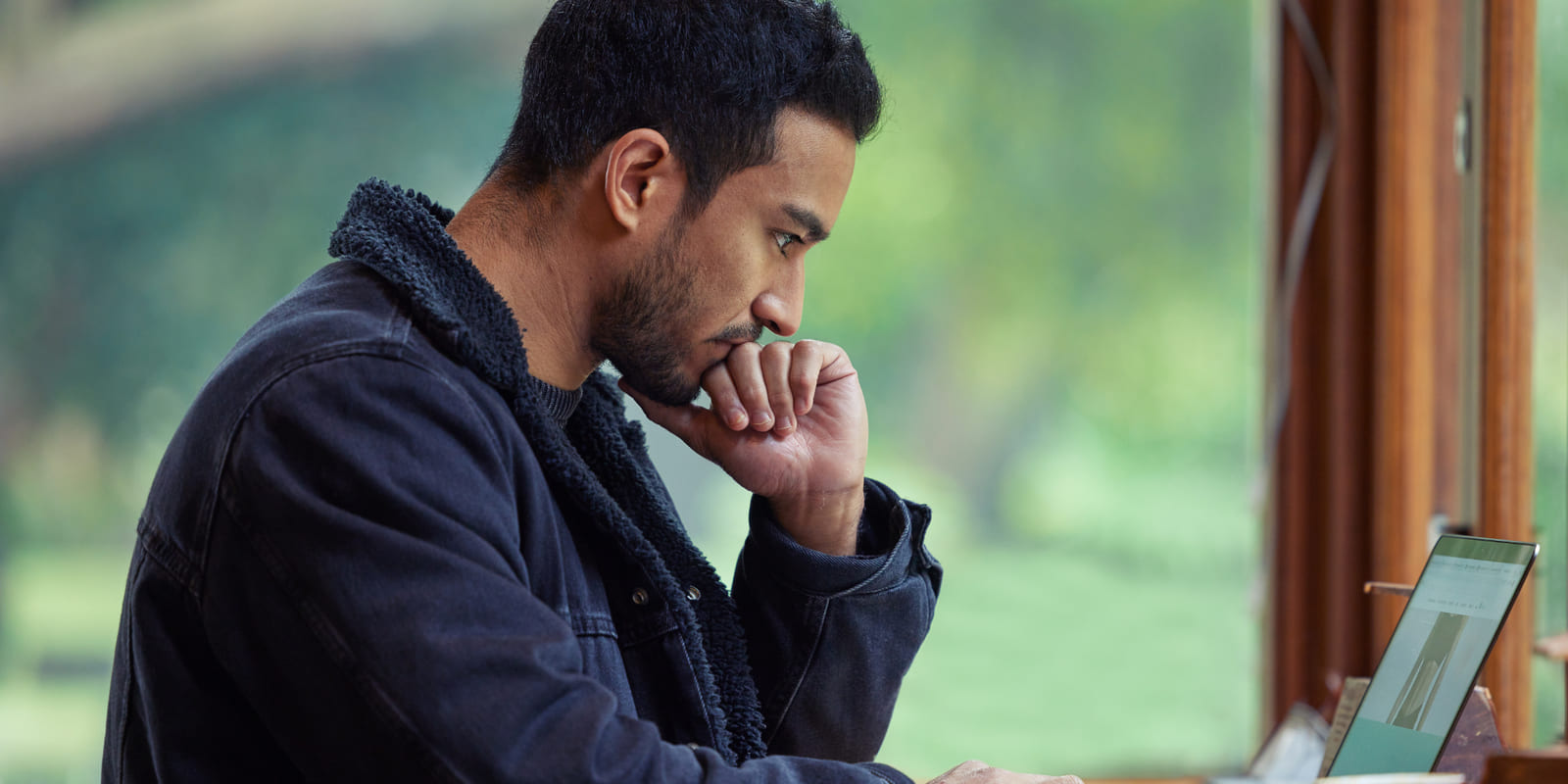 A man at a laptop by a window, hand against his mouth, taking a second look at something before he answers